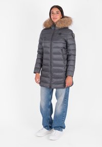 Grijze pufferjack met een gevoerde capuchon van bont, verticale gestikte design, ritszakken en een slanke pasvorm, gecombineerd met blauwe wijde jeans en witte sneakers.