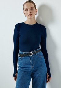 Morska majica dugih rukava koju prati bluza s visokim strukom i crni pojas sa srebrnom kopčom, na običnoj pozadini.