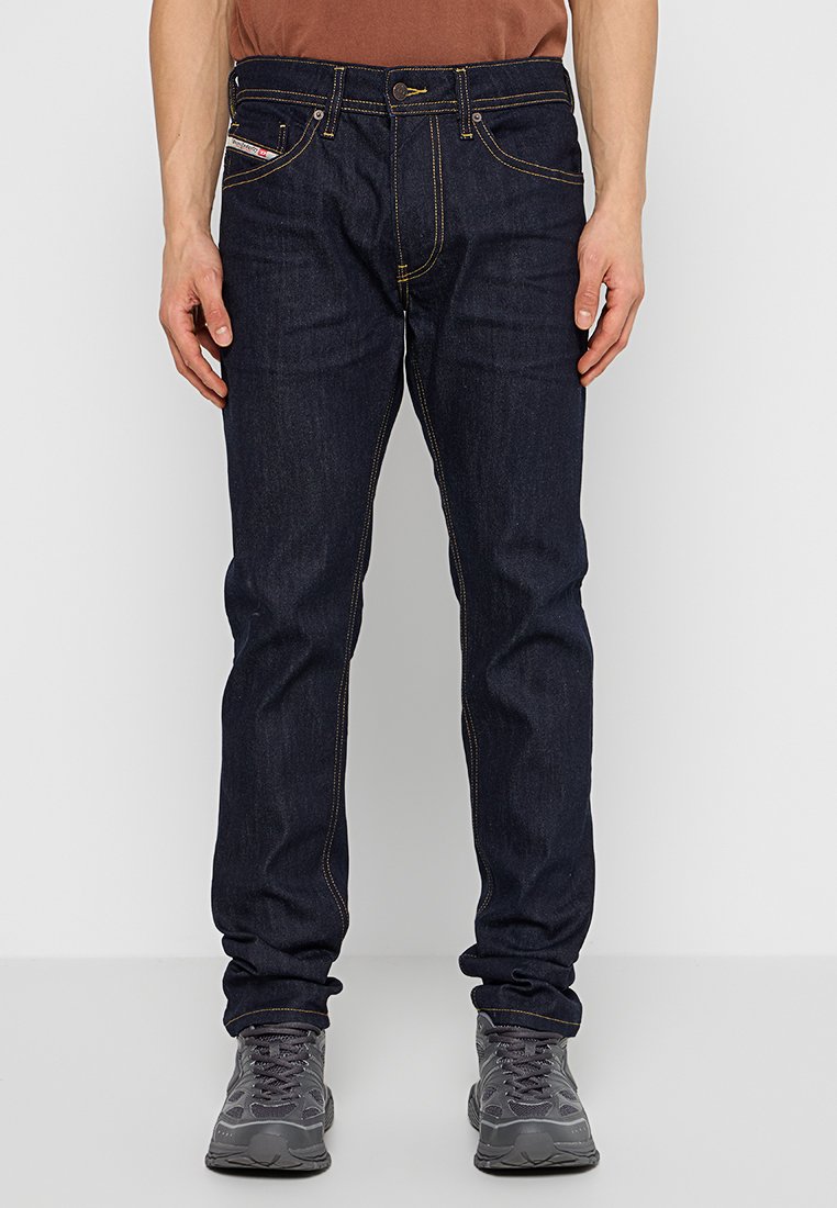 Jean en denim bleu foncé avec une coupe droite, coutures jaunes, cinq poches et fermeture à bouton. Porté avec des baskets grises.