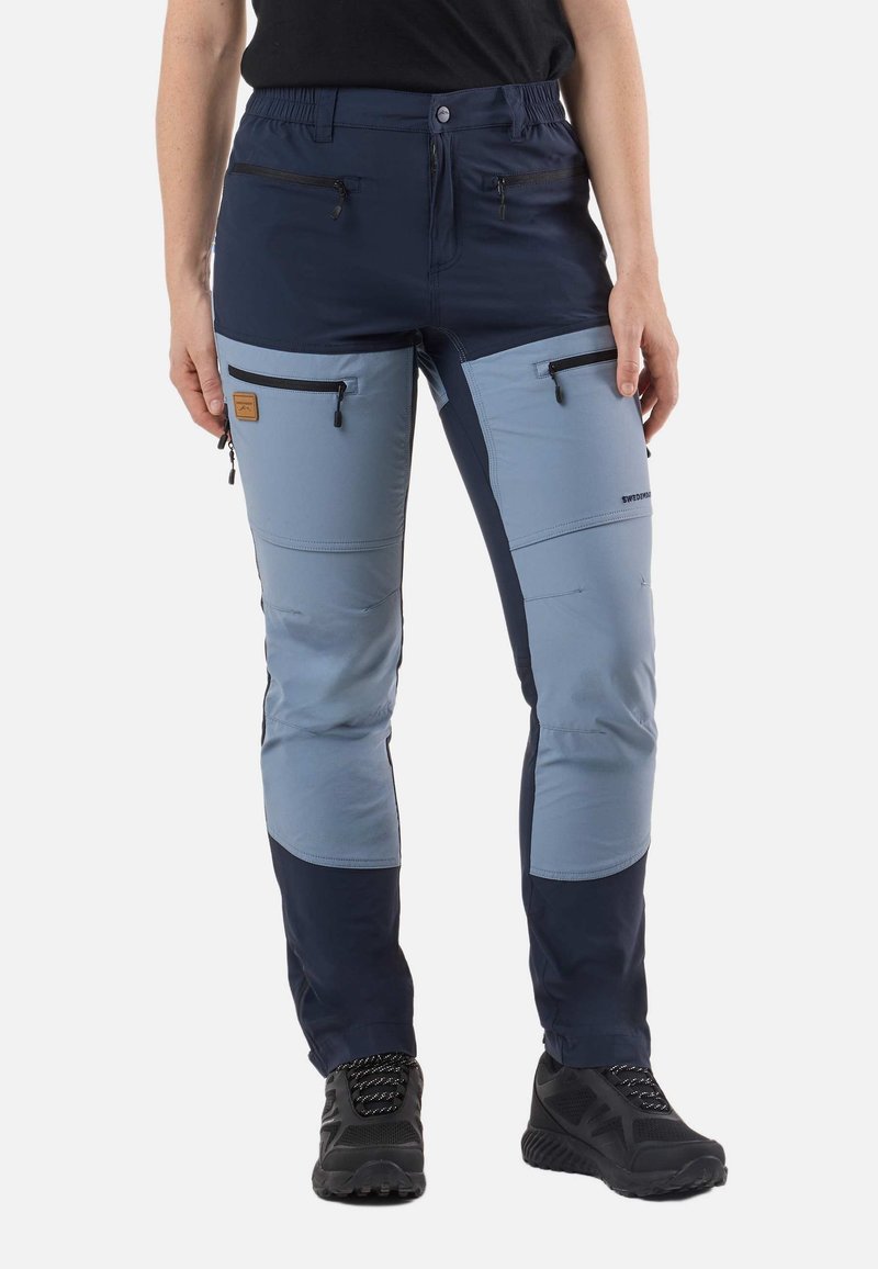 Person wearing two-tone blue outdoor pants with black zippered pockets and black hiking shoes, standing against white background.