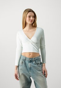 Top cropped à manches longues côtelé blanc avec un décolleté en V profond et un design enroulé, associé à un jean en denim bleu clair taille haute.