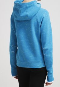 En person som bär en blå hoodie med ljusgröna ränder, långa ärmar med tumhål och svarta byxor, vänd bort.