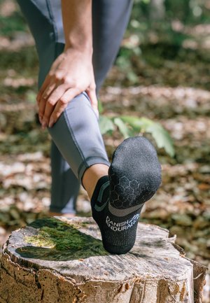 Schwarze Sportsocke mit hexagonalem Muster auf der Sohle, getragen an einem Fuß, der auf einem Baumstumpf in einer natürlichen Umgebung ruht.