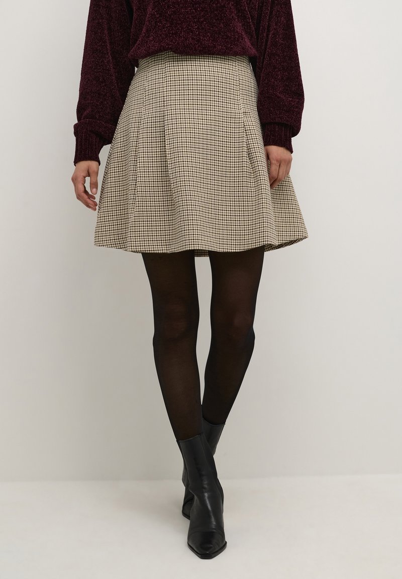 Gonna plissettata a quadri in tessuto beige e nero. Vita aderente, design svasato, lunghezza al ginocchio. Abbinata a stivali neri alla caviglia e collant.