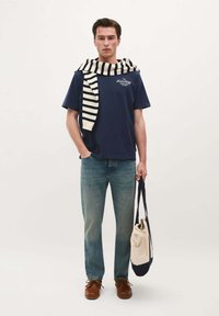 Man wearing navy t-shirt and jeans with striped sweater draped over shoulders, holding a beige and navy tote bag.