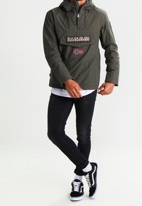 Olivengrøn pulloverjakke med hætte, frontlomme og logo-patch; parret med sorte bukser og sort/hvide sneakers. Glat tekstur.