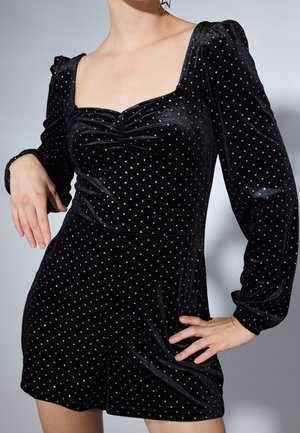 Kobieta w czarnym aksamitnym kombinezonie z bufiastymi długimi rękawami i srebrnymi groszkami, ręce opierające się na biodrze i udzie.