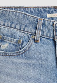 Jean en denim bleu clair en gros plan montrant une poche avant, une petite déchirure usée, des rivets argentés, un passant de ceinture et un détail de surpiqûre orange.