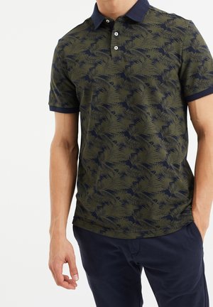 Mężczyzna w zielono-granatowej koszulce polo z nadrukiem liści palmy i granatowych spodniach, stojący z jedną ręką w kieszeni.