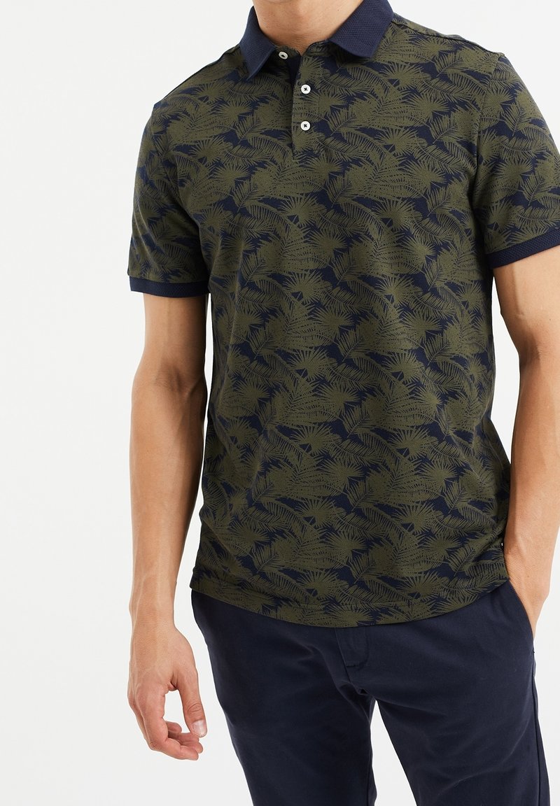 Homme portant un polo vert et marine à motif de feuilles de palmier et un pantalon marine, debout avec une main dans la poche.