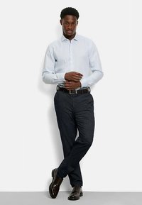 Hellblaues, langärmliges Hemd mit Knöpfen, dunkle, strukturierte Hose, brauner Ledergürtel und polierte braune Schuhe. Model posiert mit verschränkten Armen.