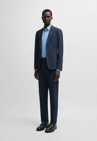 Abito blu navy con un design su misura, caratterizzato da un blazer monopetto e pantaloni coordinati. Camicia azzurra sotto. Scarpe nere.