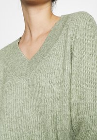 vihreä neulepaita, jossa on V-pääntie, teksturoitu kangas ja hieman väljä istuvuus. Materiaali näyttää pehmeältä ja mukavalta.