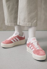 Persona con zapatillas Adidas rosa y blancas, calcetines blancos y pantalones cortos beige, de pie sobre un suelo moteado.
