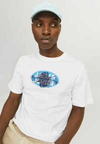 Hvid bomulds T-shirt med en blå oval grafik med teksten "JACK & JONES." Modellen bærer også en lyseblå kasket. Casual pasform, kortærmet.