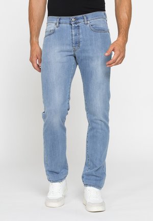 Uomo con jeans azzurri chiari e sneakers bianche in piedi davanti a uno sfondo chiaro e semplice.