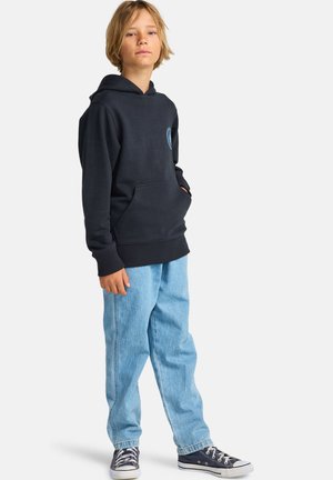 Bambino biondo in piedi con le mani nelle tasche di una felpa nera con cappuccio, indossa jeans azzurri e scarpe da ginnastica nere basse su sfondo bianco.