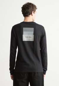 Melna garām piedurknēm T-krekls ar grafisku dizainu aizmugurē, kurā redzami slāņoti taisnstūri pelēkās toņos un zem tiem ir teksts "Calvin Klein".