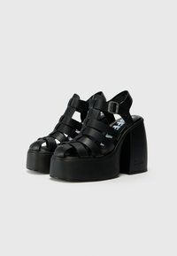 Black platform sandals featuring wide, interlaced straps and an adjustable ankle buckle. Smooth texture with a rounded toe and chunky heel.