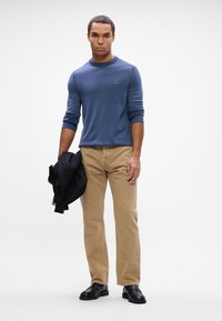 Blue crew-neck sweater, beige chinos, black shoes, and holding a black jacket. Sweater features ribbed cuffs; chinos are straight-leg.