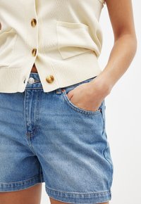 Beige stickad cardigan med guldknappar, matchad med ljusa denimshorts med en klassisk femficksdesign och subtila bleknade detaljer.