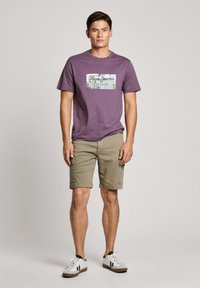Violetinis medvilninis marškinėlis su baltu grafinio spaudiniu ir tekstu, derinamas su smėliniais kargo šortais ir baltais sportbačiais su juodais akcentais.