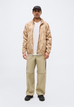 Mężczyzna w beżowej kurtce w wzory, białej koszulce, jasnobrązowych spodniach z odpinanymi nogawkami, czarnych butach i czapce stoi na jednolitym jasnym tle.