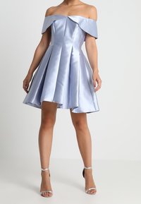 Žena v průsvitných, světle modrých saténových šatech s odhalenými rameny a sklady, v kombinaci se stříbrnými sandály na vysokém podpatku, stojící na bílém pozadí.