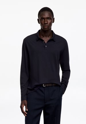 Homme debout portant un polo noir à manches longues et un pantalon foncé, avec une main dans la poche sur un fond blanc uni.
