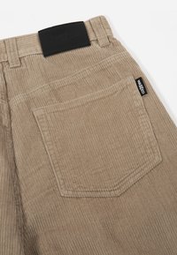 Beige cordfløyel-bukser med vertikal ribbestruktur, en svart midjebåndslabel og en sydd baklomme med label.