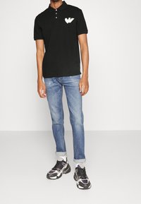 Svart pikétröja med en vit grafisk logo på vänster bröst, kombinerad med ljusblå denimjeans och svarta sneakers med grå detaljer.