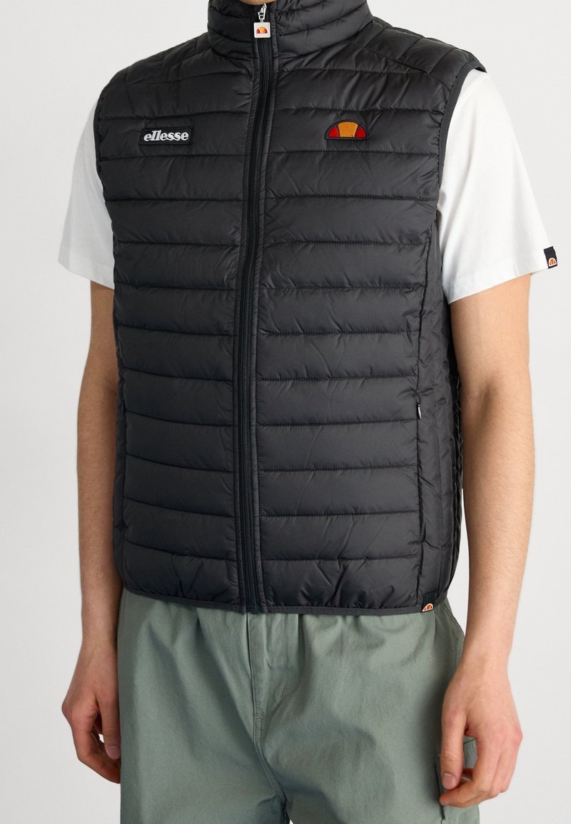 Homme portant un gilet matelassé noir sans manches sur une chemise blanche à manches courtes et un pantalon vert, gilet zippé avec les logos de la marque visibles.