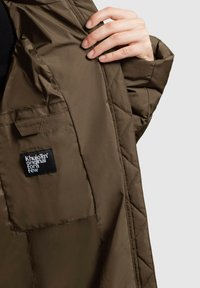 Käsi hoiab avatud pruuni vatitud jope, mille seest on nähtav tasku musta sildiga, millel on kirjas "Khujo™ originaal mõnele."