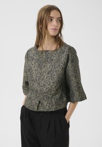 Bluză cu mânecă scurtă dintr-un material texturat de culoare verde închis, cu accente aurii, având un decolteu rotund și detaliu peplum. Pantaloni pliți negri.