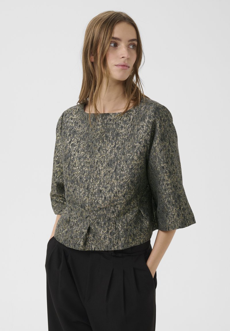 Bluză cu mânecă scurtă dintr-un material texturat de culoare verde închis, cu accente aurii, având un decolteu rotund și detaliu peplum. Pantaloni pliți negri.