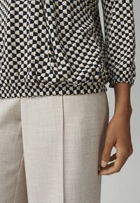 Gemusterte Bluse mit schwarzen und cremefarbenen geometrischen Formen, an der Taille gerafft, kombiniert mit hellbeigen strukturierten Hosen.