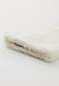 Material fuzzy di colore crema con una superficie testurizzata e un accento in tessuto bianco. L'estremità è ripiegata, rivelando un bordo liscio.