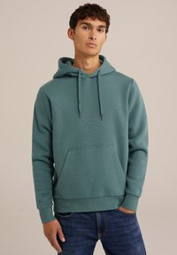 Hanorac cu glugă de culoare teal, fabricat dintr-o țesătură moale. Include un buzunar tip cangur și manșete riglate. Poartă cu blugi închiși la culoare.