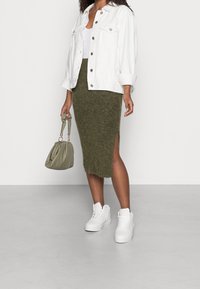 Vit denimjacka, olivgrön stickad kjol med sidoklyfta, vita högstövlade sneakers och en mjuk olivgrön handväska.