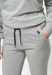Lichtgrijze sweatpants gemaakt van zachte stof, met een marineblauwe trekkoord, zijzakken en een klein marineblauw geborduurd logo op de linkse zak.