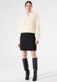 Suéter crema acanalado con cuello de media cremallera, combinado con una falda corta negra con una abertura lateral y botas de cuero negras hasta la rodilla.