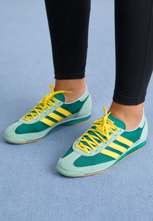 Henkilö, joka käyttää vihreitä ja vaaleansinisiä Adidas-urheilujalkineita, joissa on keltaiset raidat ja nauhat, seisoo vaaleansinisellä matolla.