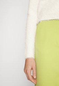 Haut blanc à manches longues, duveteux avec une texture douce, associé à une jupe verte vive ajustée. Gros plan montrant la manche et la position de la main.