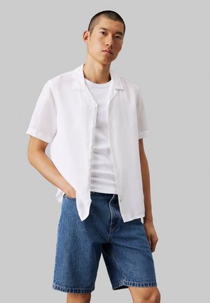 Camisa de botones de manga corta blanca en un material ligero, superpuesta a una camiseta sin mangas blanca de canalé. Combinada con pantalones cortos de mezclilla azul.