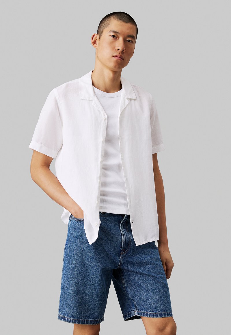 Camisa de botones de manga corta blanca en un material ligero, superpuesta a una camiseta sin mangas blanca de canalé. Combinada con pantalones cortos de mezclilla azul.