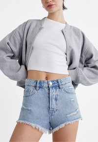 Mujer vistiendo pantalones cortos de mezclilla desgastados de color azul claro, top corto blanco y chaqueta bomber gris con botones, con las manos en las caderas.
