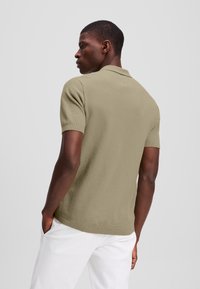 Khaki polo tričko s krátkym rukávom, textúrovaná látka a malý logo detail na chrbte. V kombinácii s bielymi nohavicami.