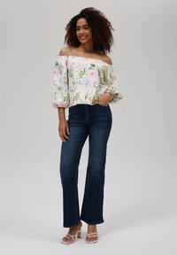 Off-shoulder blomstret bluse i hvit med rosa og lilla blomster; paret med mørkeblå flytende jeans og hvite hæl sandaler.