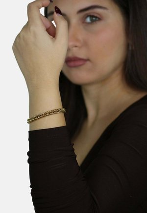 Bracciale d'oro con catene intrecciate, indossato su un polso con una maglietta a maniche lunghe aderente. Texture liscia e finitura lucida.