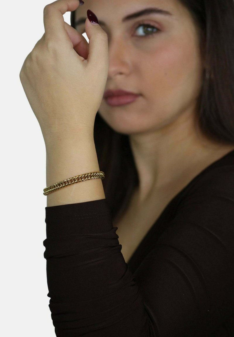 Bracciale d'oro con catene intrecciate, indossato su un polso con una maglietta a maniche lunghe aderente. Texture liscia e finitura lucida.